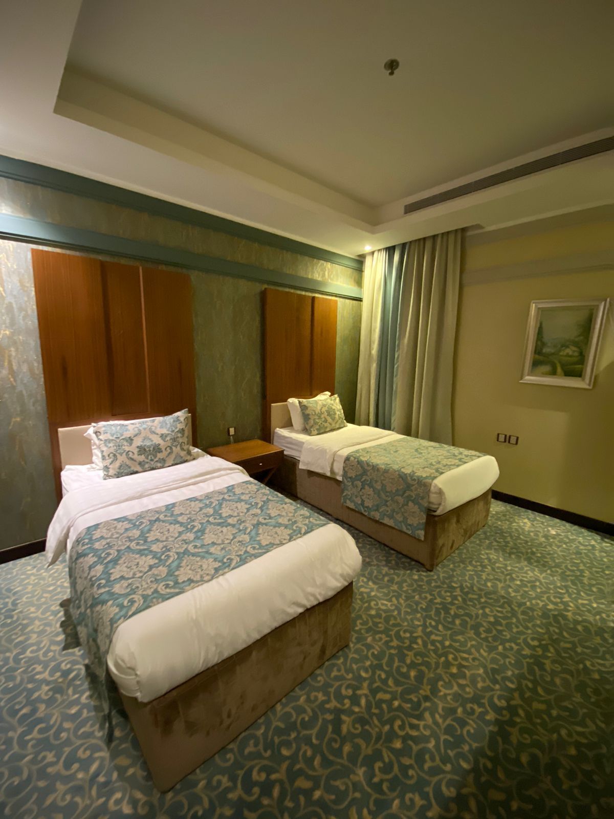 A hotel room with two beds, a wooden wardrobe, a nightstand, and a painting on the wall.