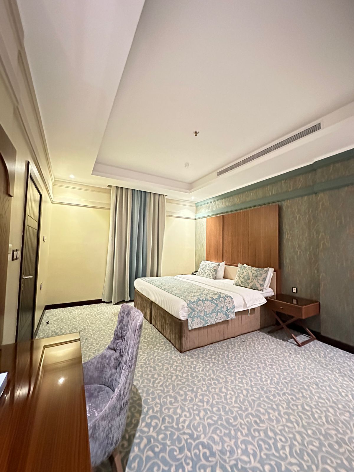A hotel room with a large bed, a wooden nightstand, a chair, and a wardrobe.