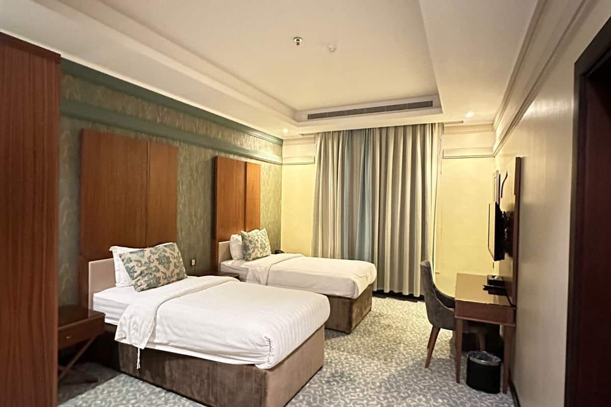 Ein Hotelzimmer mit zwei Betten, einem Schreibtisch, einem Sessel, einem Fernseher und einem gemusterten Teppich.