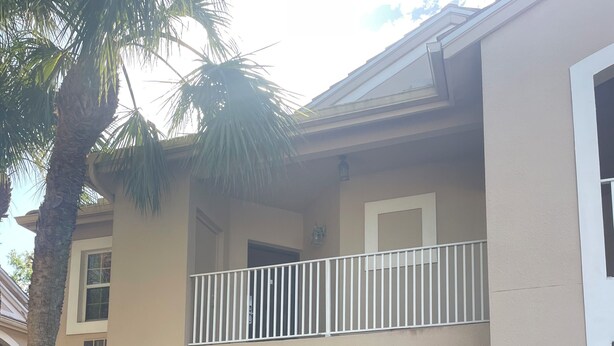 Un edificio de dos pisos con un balcón, rodeado de plantas diversas y una palmera.