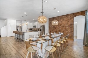A spacious kitchen with modern appliances, a dining area with a long table set for six, and a wooden accent wall. - Arcade, Waterpark, Theater, LEGO Room, Pool & Spa, Dual 86” TVs: Paradise Palms (Davenport)