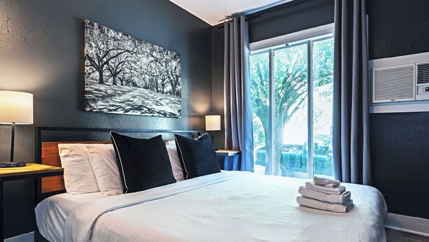 A hotel room with a large bed, two bedside tables, a wall-mounted air conditioner, and a window with curtains.
