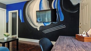 A modern living room with a large abstract mural, a flat-screen TV mounted on the wall, a wooden dining table, and dark wooden flooring.