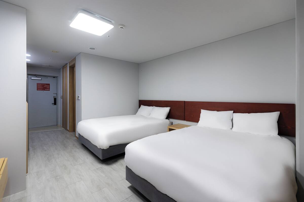 Habitación de hotel con dos camas, un cabecero de madera, una mesita de noche y una puerta que da a otra habitación.
