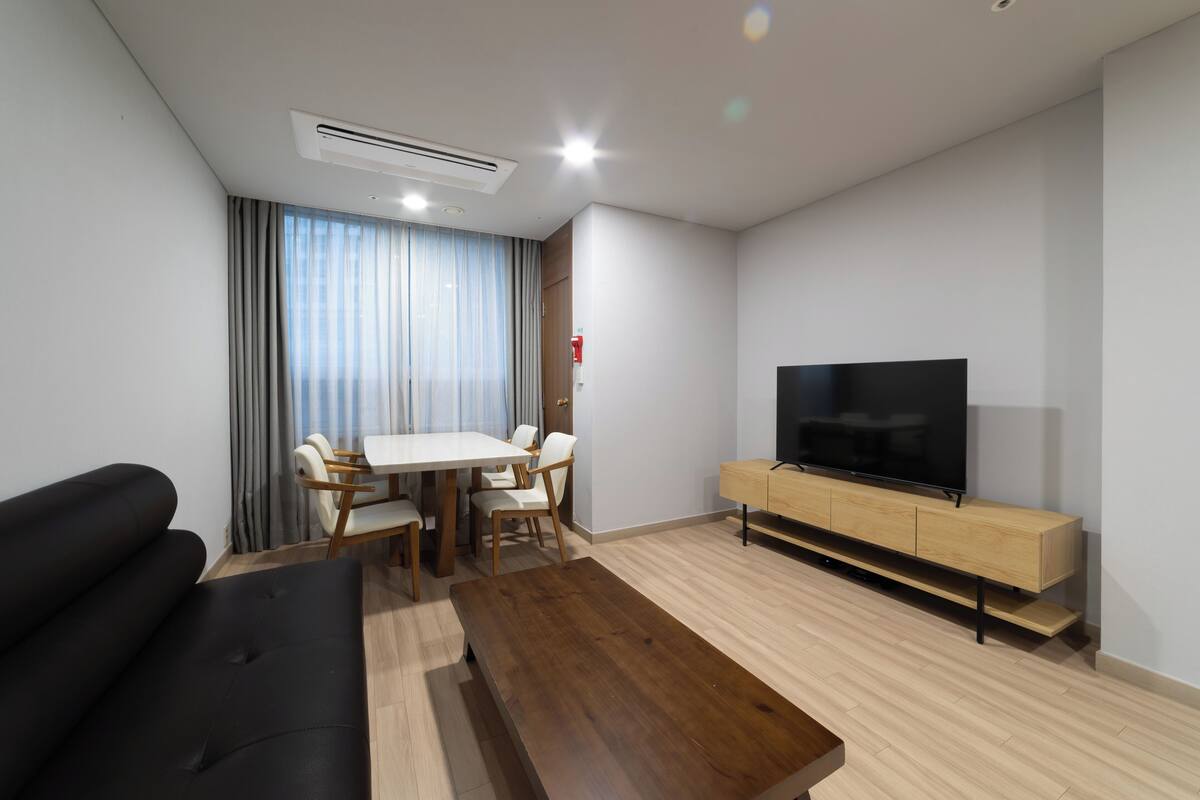 Una sala moderna con un sofá, una mesa de centro de madera, un televisor sobre un mueble de madera, una mesa de comedor con sillas y una ventana con cortinas.
