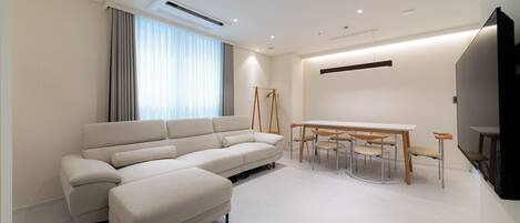 A modern living room with a large L-shaped sofa, a dining table with chairs, and a flat-screen TV mounted on the wall.