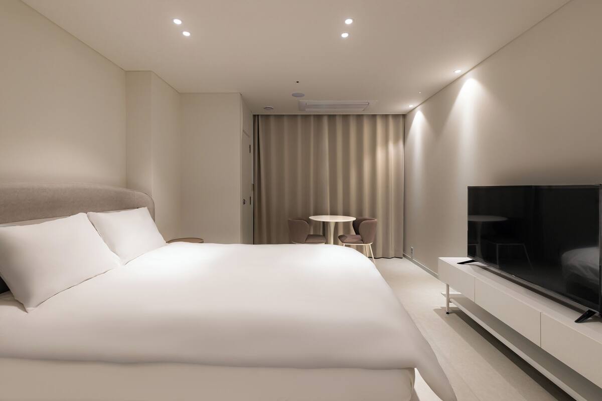 Una habitación de hotel moderna con una cama grande, un televisor de pantalla plana, una mesita redonda y una zona de estar con cortinas.