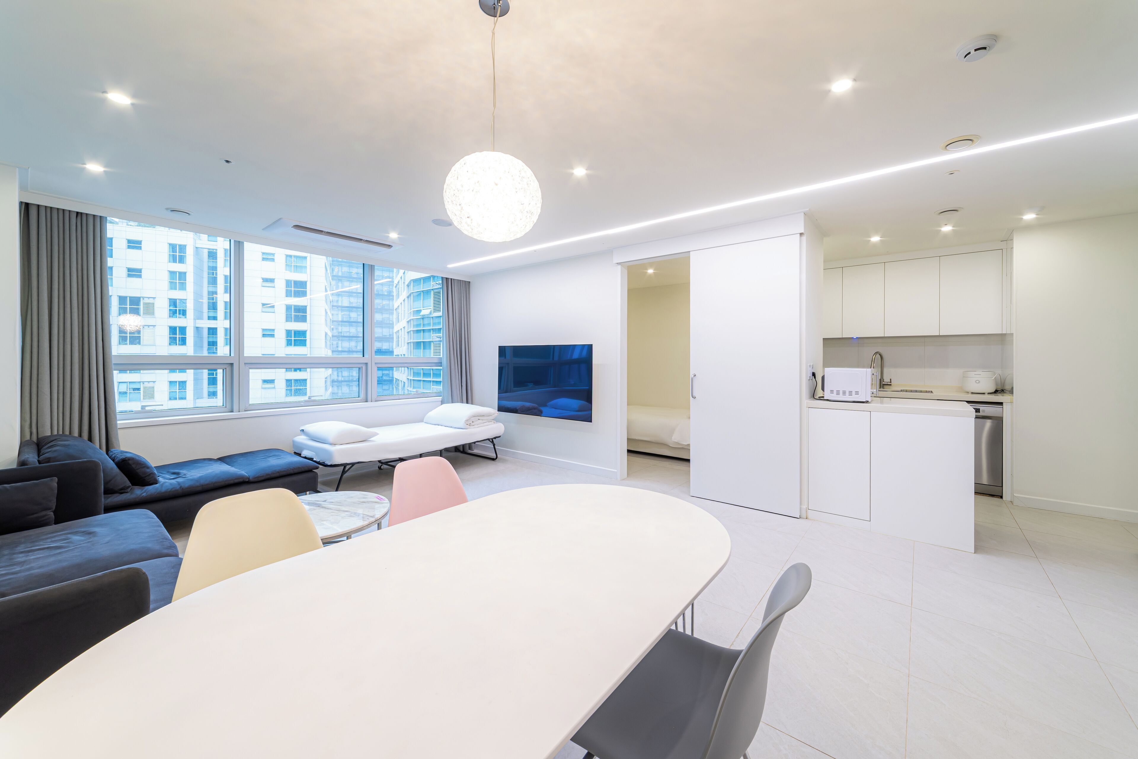 A modern hotel room with a large window, a dining area, a sofa, a TV, and a kitchenette.