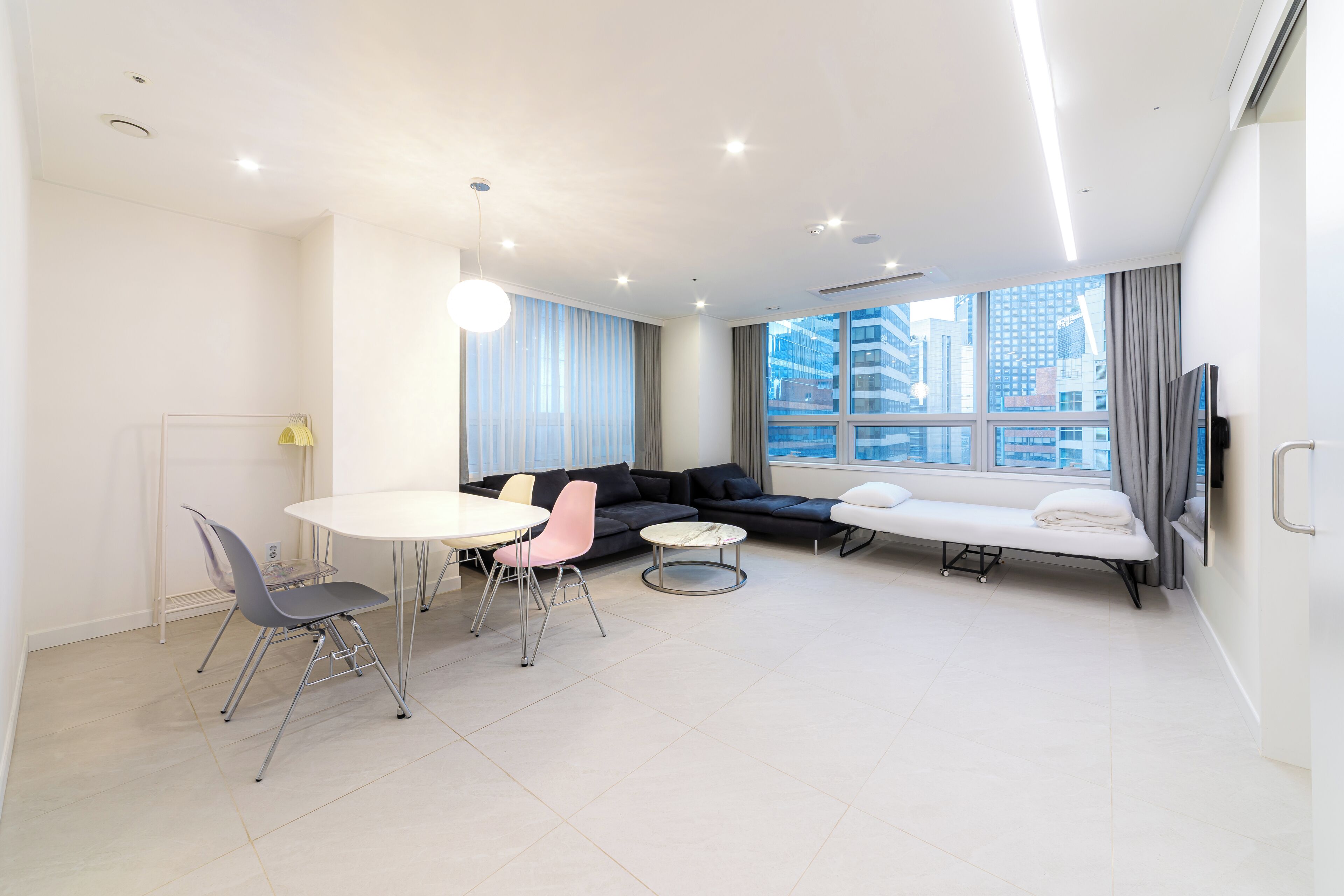 A modern living room with a sofa, a dining table with chairs, a small round table, and a large window with a city view.