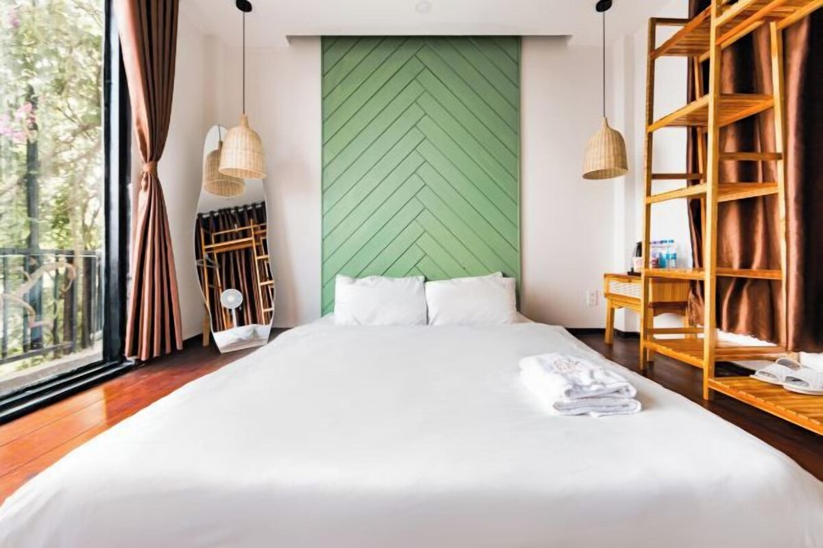 A hotel room with a large bed, a green herringbone-patterned wall, wooden shelves, and a view of the outdoors.