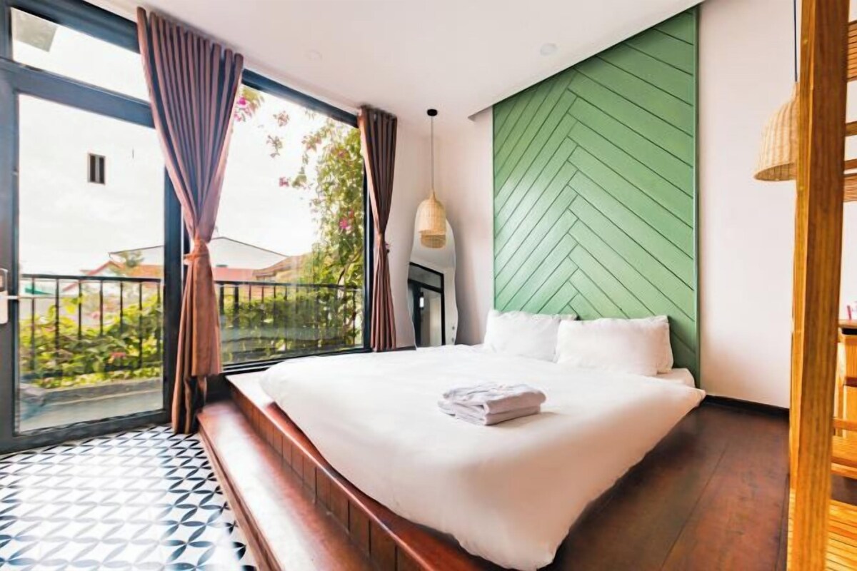 A bedroom with a large bed, a green accent wall, and a view of greenery outside.