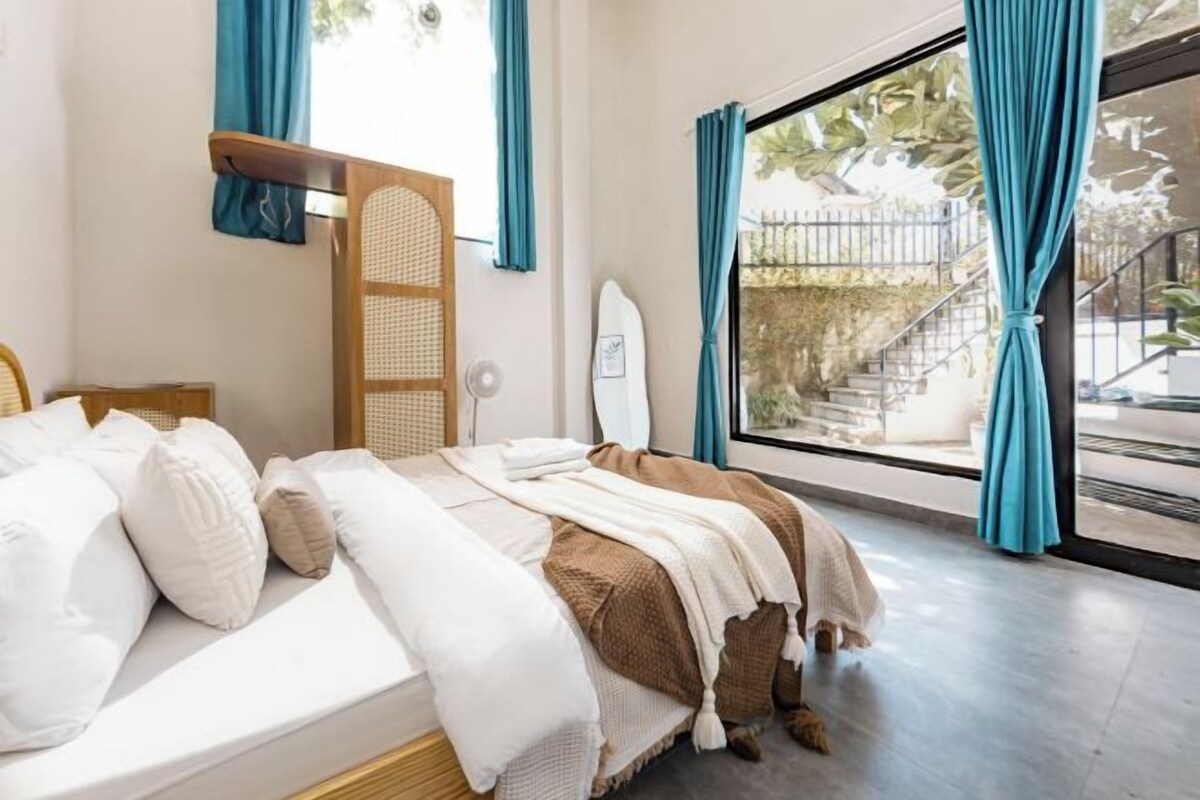 A bedroom with a bed, a window with curtains, a surfboard, and a staircase visible outside.
