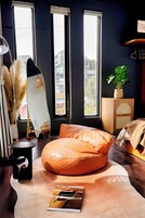 Um quarto com uma janela grande, um puff, uma mesinha com luminária e uma planta em vaso.