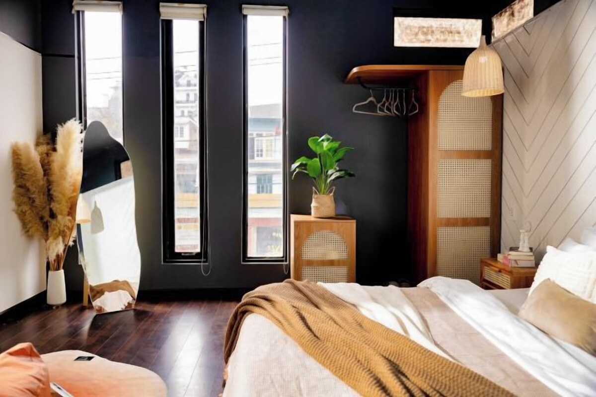 A modern bedroom with a large bed, a side table with a plant, a hanging light fixture, and a view of the city through large windows.