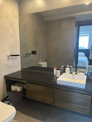 A modern bathroom with a dark marble countertop, a white sink, and a large mirror. There are toiletries on the counter and a toilet paper holder below. - Torres Evolution - Charming 1-bedroom apartment in fabulous Santa Cruz (Santa Cruz de la Sierra)
