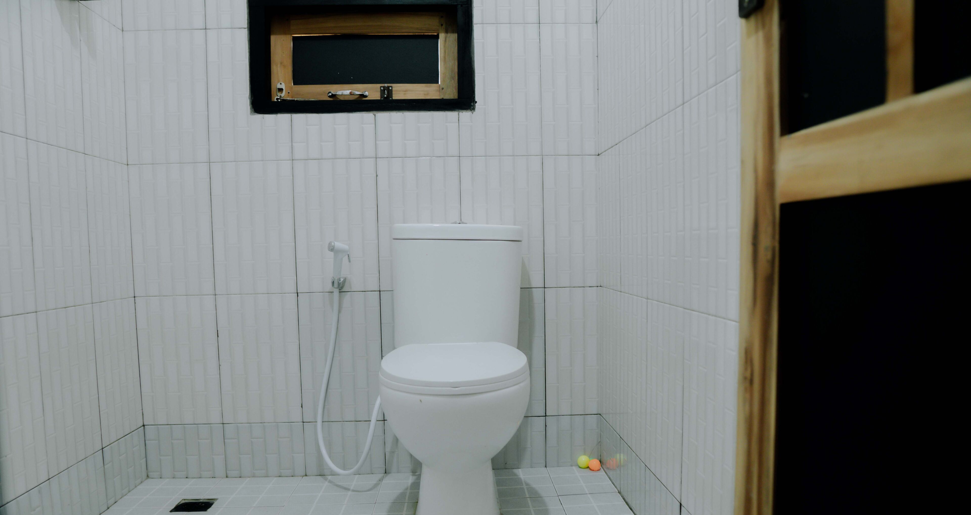 A small, tiled bathroom with a white toilet, a showerhead, and a small wooden shelf above the toilet.
