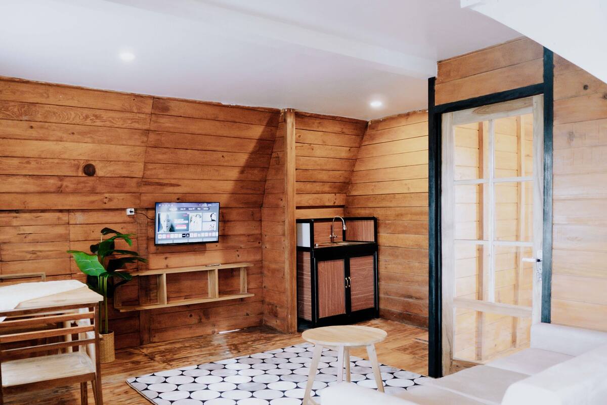 A cozy room with wooden walls, a TV mounted on the wall, a small table, a chair, and a sofa.