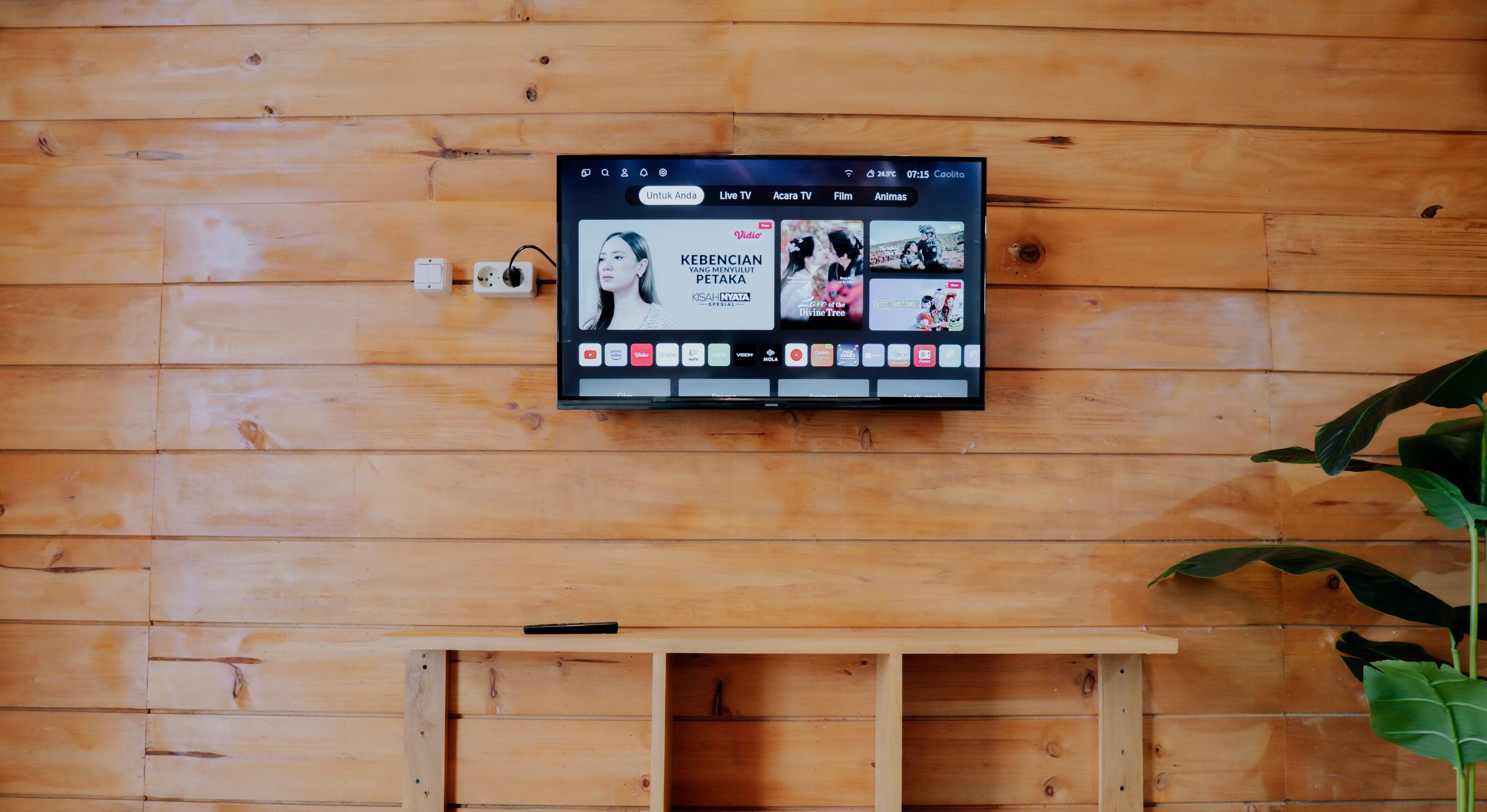 A wooden wall with a mounted TV displaying a movie titled "KEBENCIAN YANG MENYULUT PETAKA" and a wooden bench below.