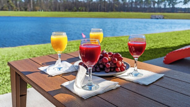 A wooden table with three glasses of juice and a plate of grapes, set against a lakeside view. - Central location Modern Lakefront 2 king-Suite 3 Bedroom 3 Full Bathroom (Myrtle Beach)