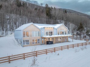 A two-story wooden cabin with a covered porch, surrounded by a wooden fence and snow-covered ground.