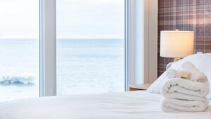 Quarto de hotel com uma janela ampla que oferece vista para o mar, cama bem arrumada com lençóis brancos e uma luminária na mesa de cabeceira.