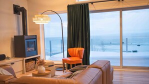 Uma sala de estar com sofá, poltrona, televisão, luminária de chão e vista para uma paisagem nevada através de grandes janelas.
