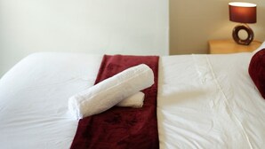 Een net opgemaakt bed met een witte deken en een rode plaid, een schilderij van mensen die in de regen lopen aan de muur, en een bedlamp met een bruine kap.