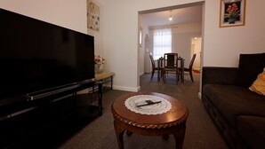Een woonkamer met een flatscreen televisie, een ronde houten salontafel met een kantekenbordje, een donkergrijze bank en een eethoek met een tafel en stoelen.