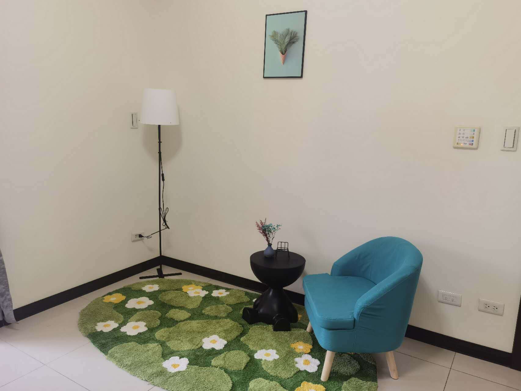 Une pièce d’angle avec un fauteuil turquoise, une petite table d’appoint noire sur laquelle est posé un vase, un lampadaire et un tapis à motifs floraux verts.