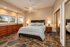 Une chambre à coucher avec un grand lit, deux tables de chevet avec des lampes, une commode en bois, un ventilateur de plafond et un grand miroir.