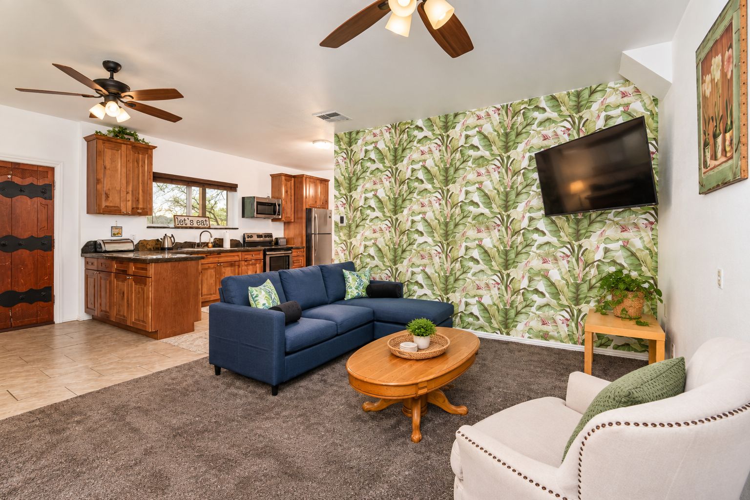 A living room with a sectional sofa, a coffee table, a TV mounted on the wall, and a kitchen in the background.