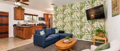 A living room with a sectional sofa, a coffee table, a TV mounted on the wall, and a kitchen in the background.