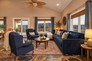 Ein geräumiges Wohnzimmer mit einer großen Couch, Sesseln, einem Couchtisch und Blick ins Freie durch große Fenster.