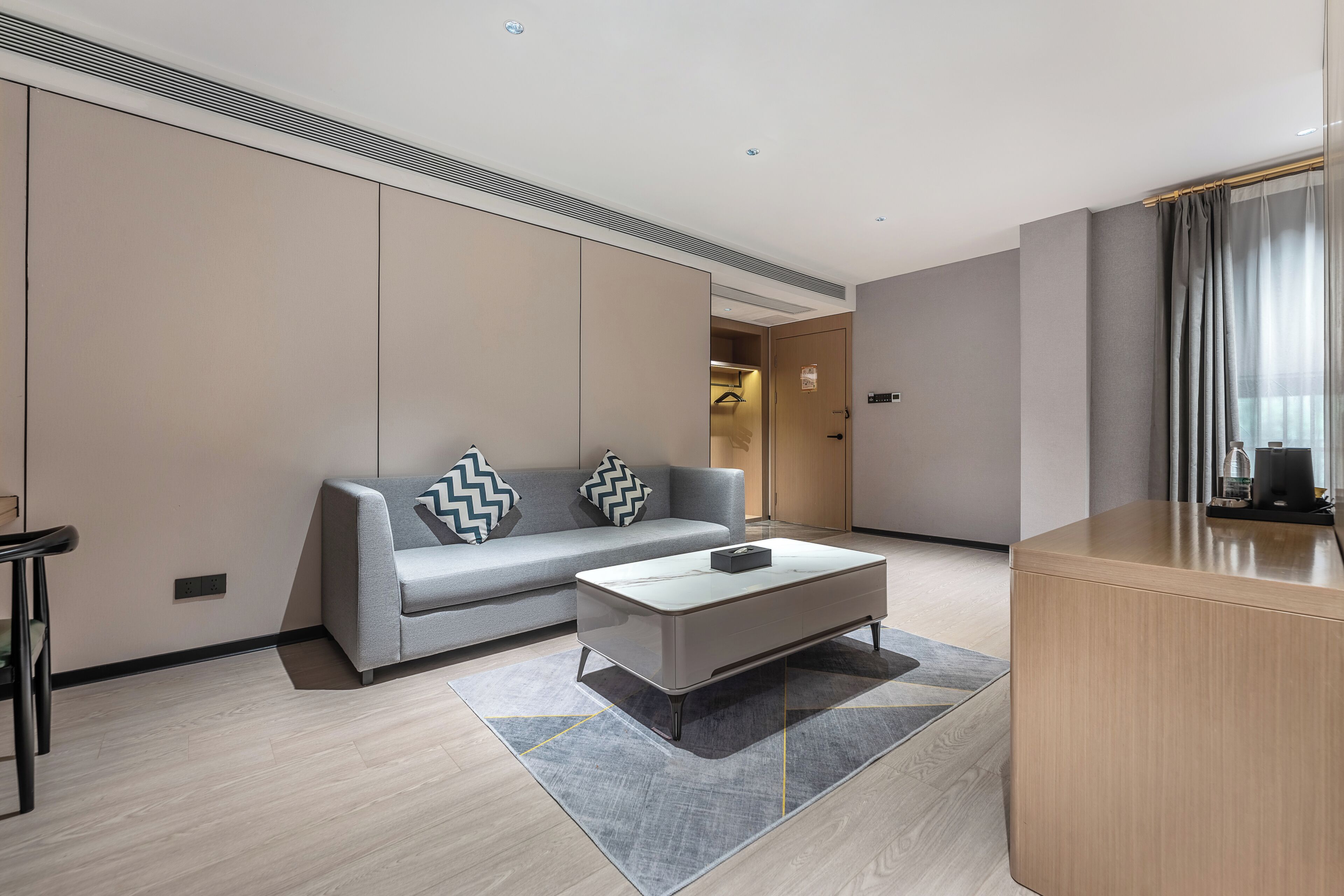 Una habitación de hotel moderna con un sofá gris, una pequeña mesa de centro, un escritorio de madera y una alfombra gris.