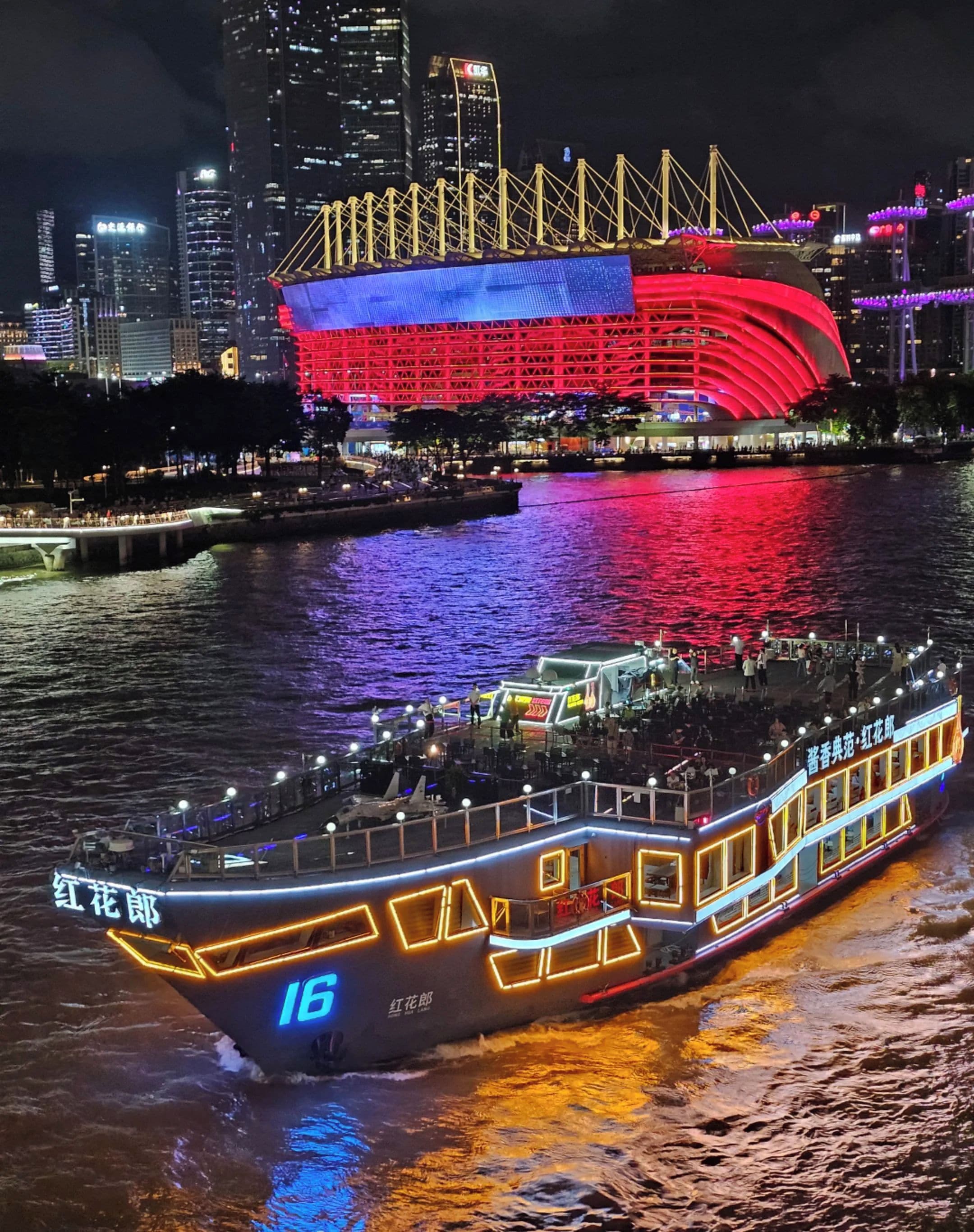 A riverboat with illuminated windows and signage, docked near a city skyline with a prominent red-lit stadium in the background.
