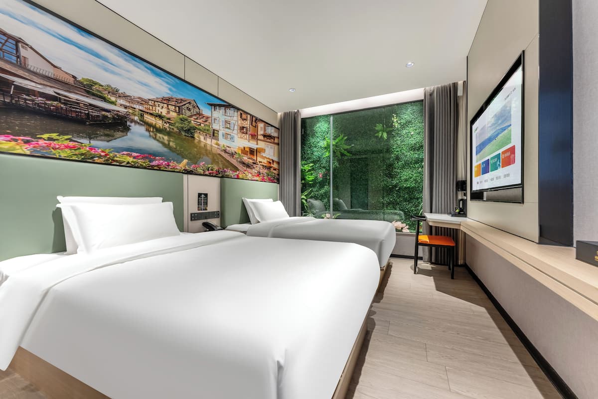 Una habitación de hotel moderna con dos camas, un ventanal con vistas a zonas verdes, un cuadro de un pueblo en la pared y un televisor de pantalla plana en la pared.