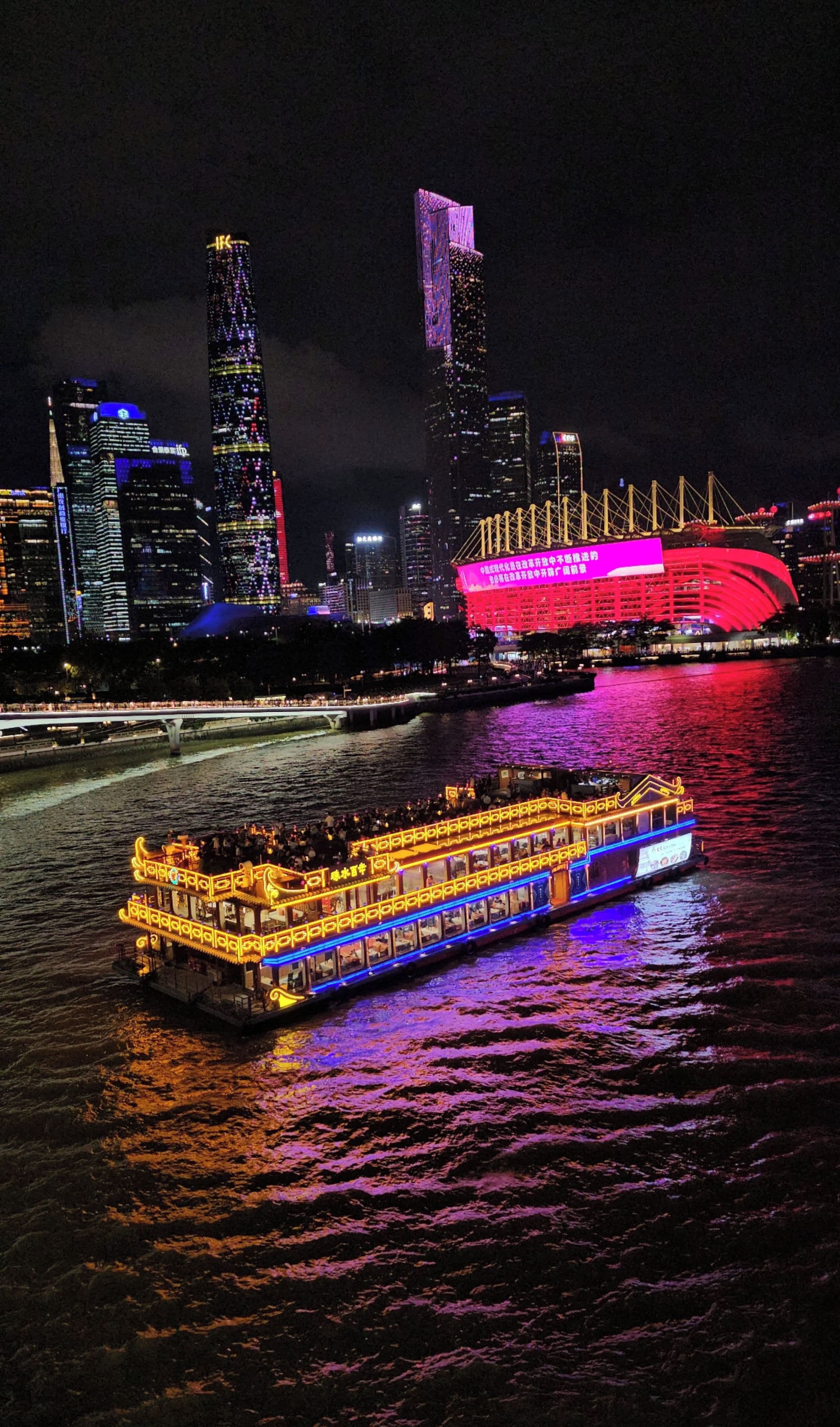 A nighttime cityscape with illuminated buildings and a river with a boat.
