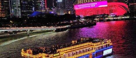 A nighttime cityscape with illuminated buildings and a river with a boat.