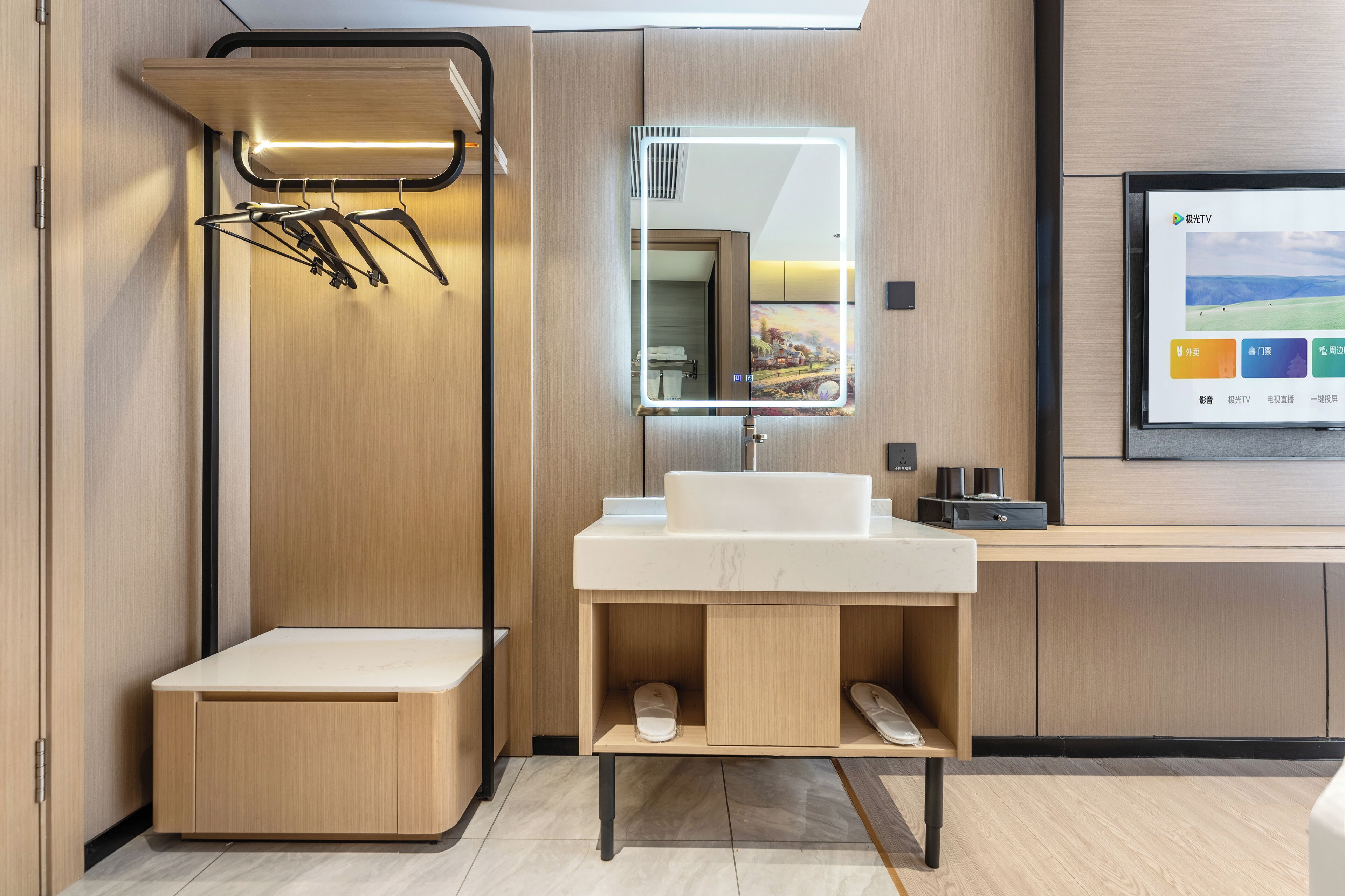 A modern bathroom with a wooden vanity, a mirror with integrated lighting, a towel rack, and a digital screen displaying a landscape.