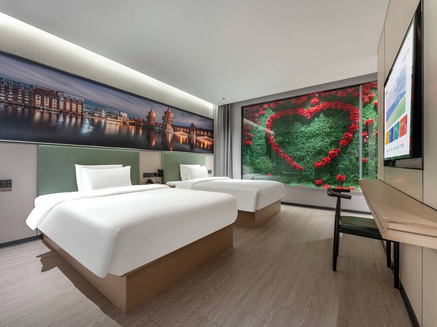 Una habitación de hotel con dos camas, un ventanal con decoración floral en forma de corazón y un mural con un paisaje urbano en la pared.