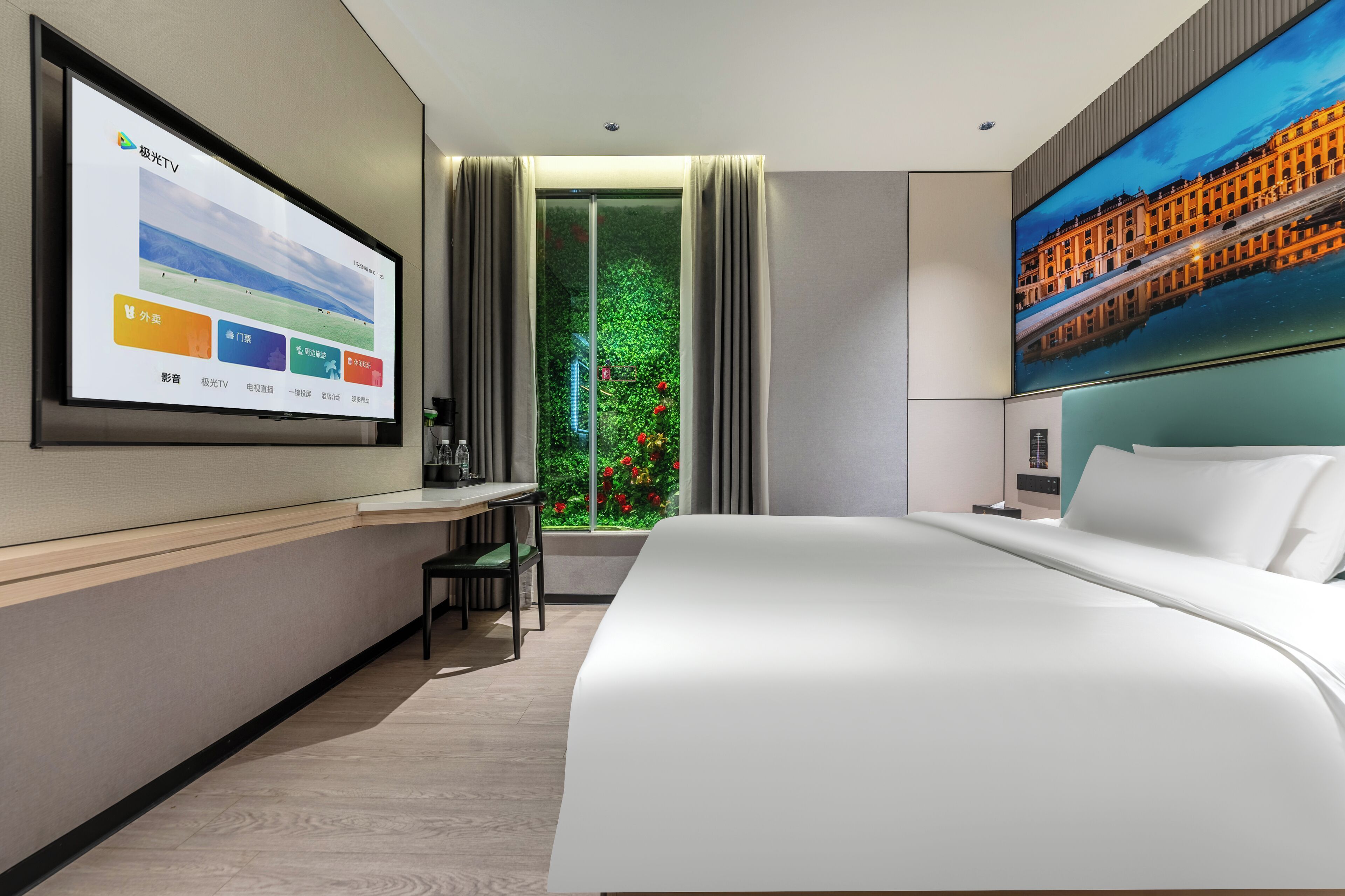 A modern hotel room with a large bed, a desk with a chair, a TV mounted on the wall, and a window with a view of greenery and a building.