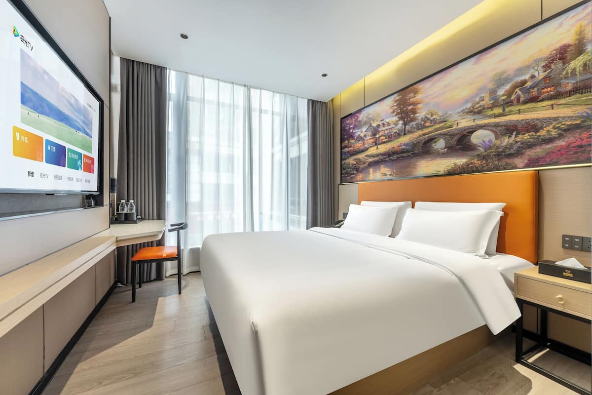 Una habitación de hotel moderna con una cama grande, un escritorio con una computadora y un televisor mostrando una interfaz colorida.