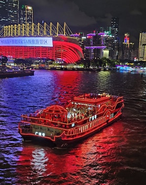 A river with a brightly lit boat and a large red building with a sign in the background.