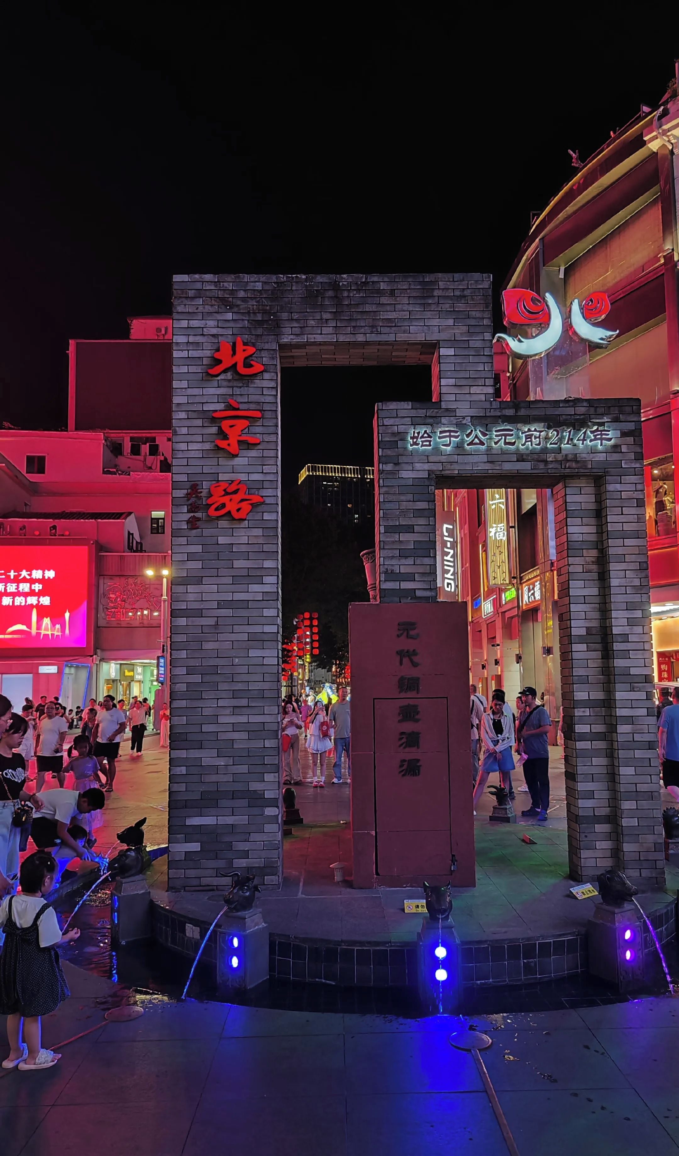 A brick entrance with illuminated signage, a water feature, and a crowd of people.