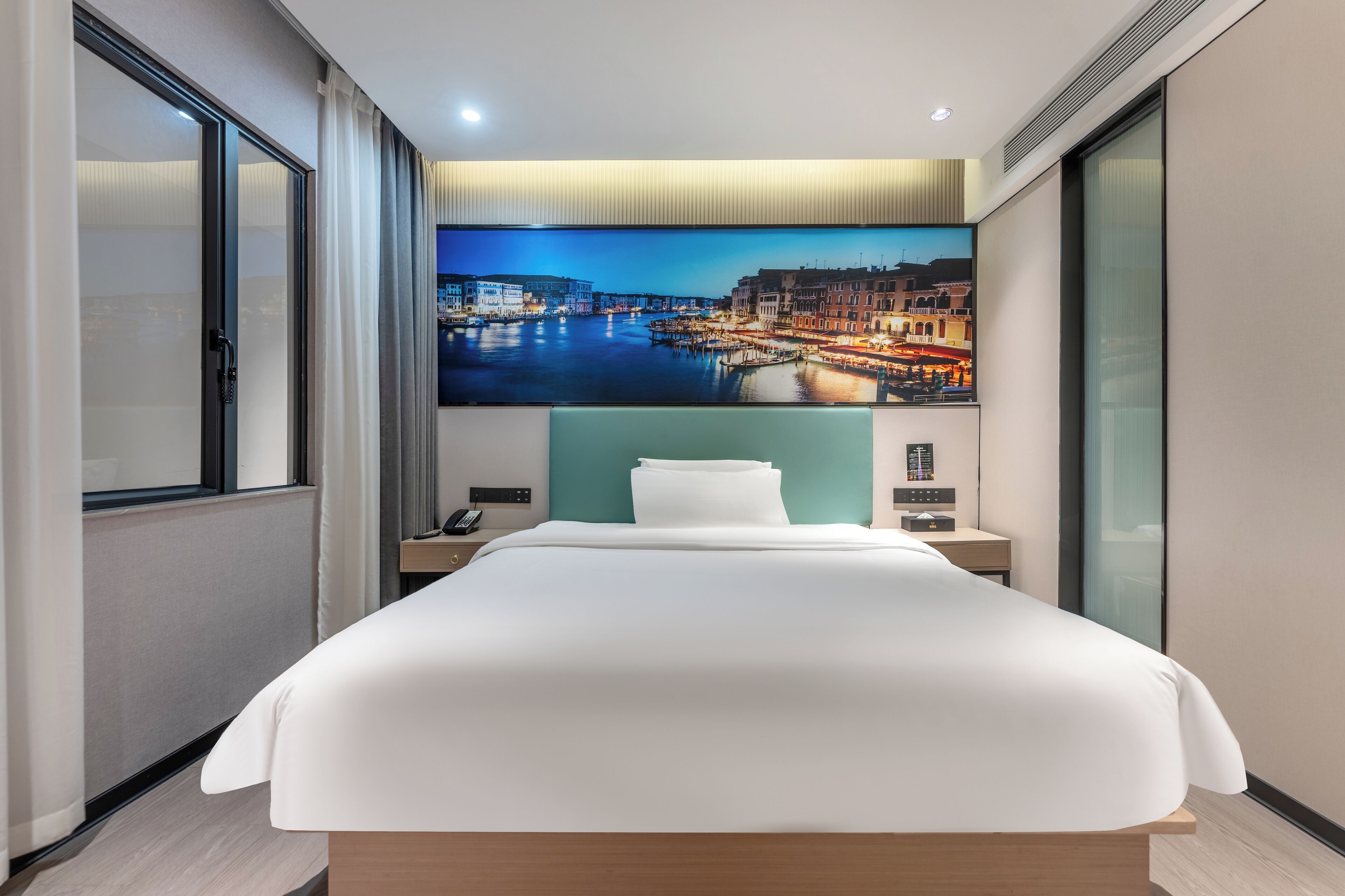 Una habitación de hotel moderna con una cama grande, un televisor de pantalla plana montado en la pared, una mesita de noche con una lámpara y una ventana con cortinas.