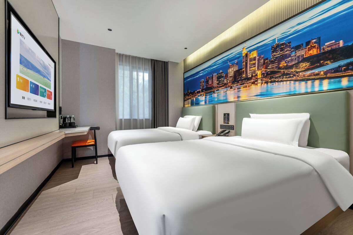 Habitación de hotel con dos camas, un escritorio, una silla, un televisor y un cuadro grande con una vista urbana.