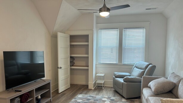 A living room with a flat-screen TV, a grey sofa, a white cabinet, and a ceiling fan. - Main Stay on Market Unit 3 -Healthcare and Military Friendly (Mascoutah)