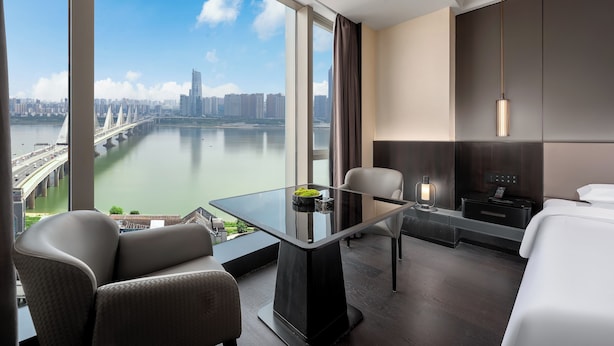 Une chambre d’hôtel moderne avec une table en verre, deux chaises, un lit, une table de chevet avec une lampe, et une vue sur le paysage urbain et la rivière.