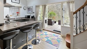A modern kitchen with a central island, bar stools, and a dining area with a view to a garden. - The Cottage at Woodlands (Lynton)