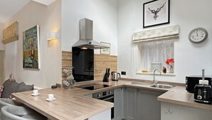 A modern kitchen with a central island, a built-in oven, and a sink. There are bar stools and a window with a view of the outdoors. - The Cottage at Woodlands (Lynton)